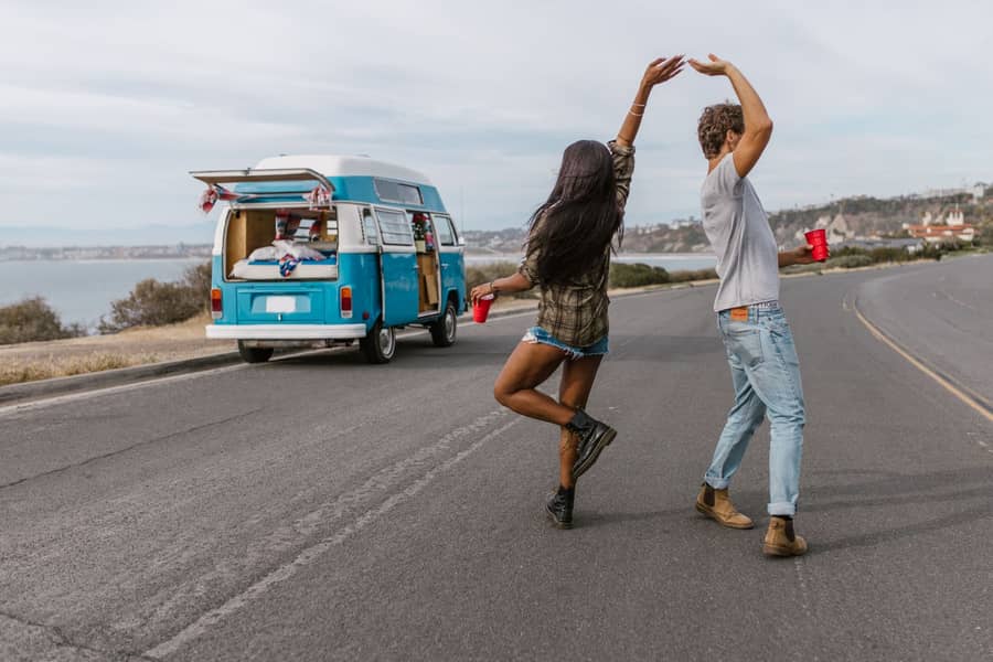 Two backpackers dancing on a coastal road next to their camper van